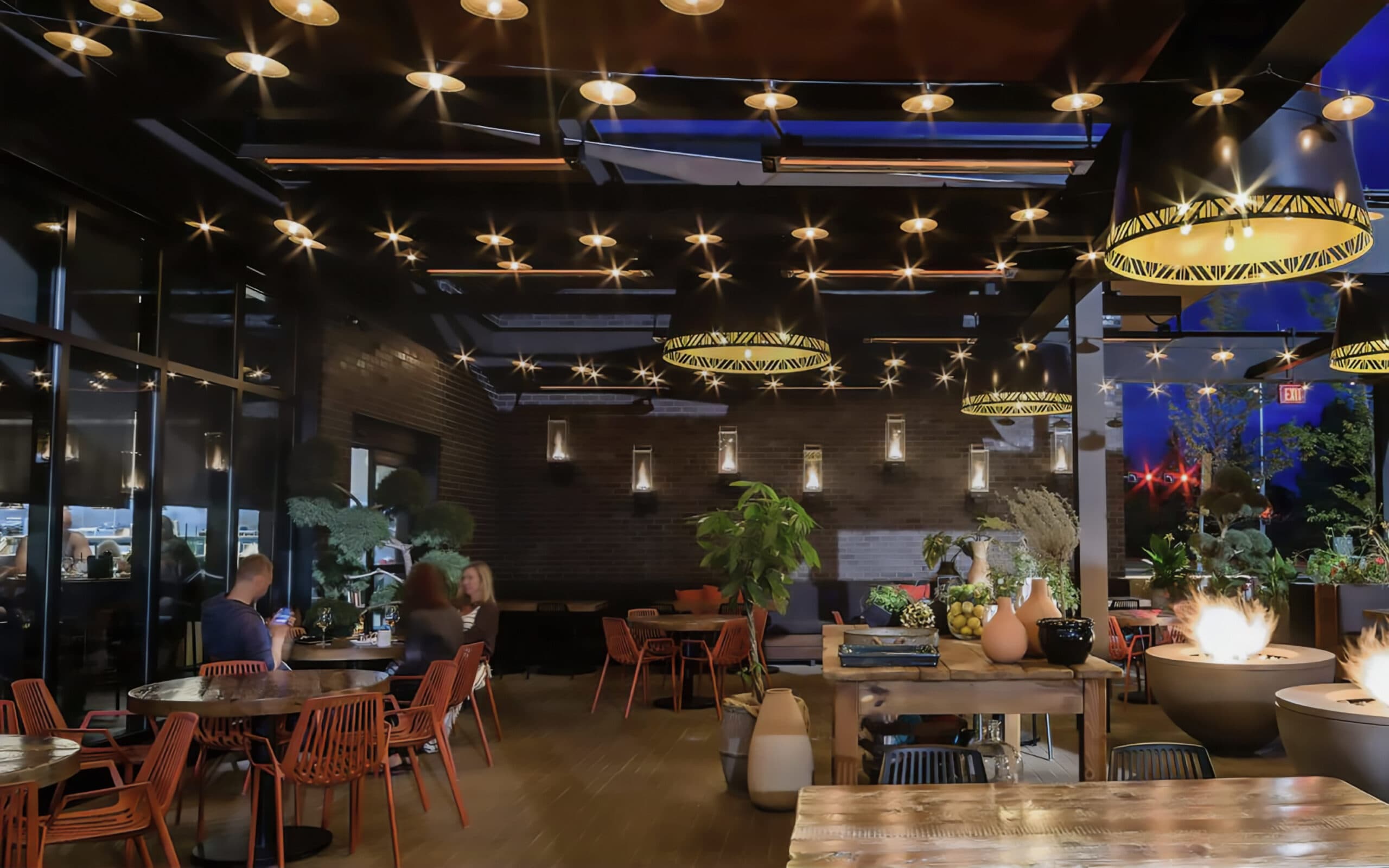 Patio with ambient lighting, wooden tables, decorative plants, string lights and large windows looking into the restaurant.