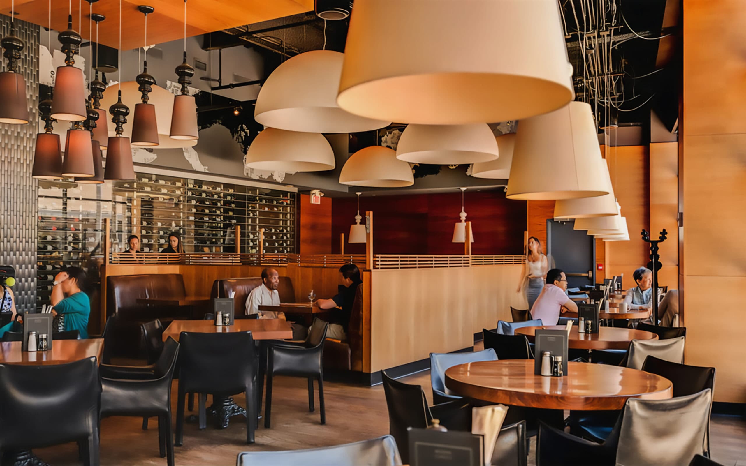 Busy restaurant interior with warm natural light, large light fixtures, wooden tables and black chairs, with a long wine wall in the background.